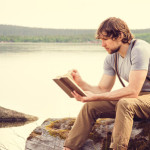 55633056 - young man reading book outdoor with scandinavian lake on background education and lifestyle travel concept